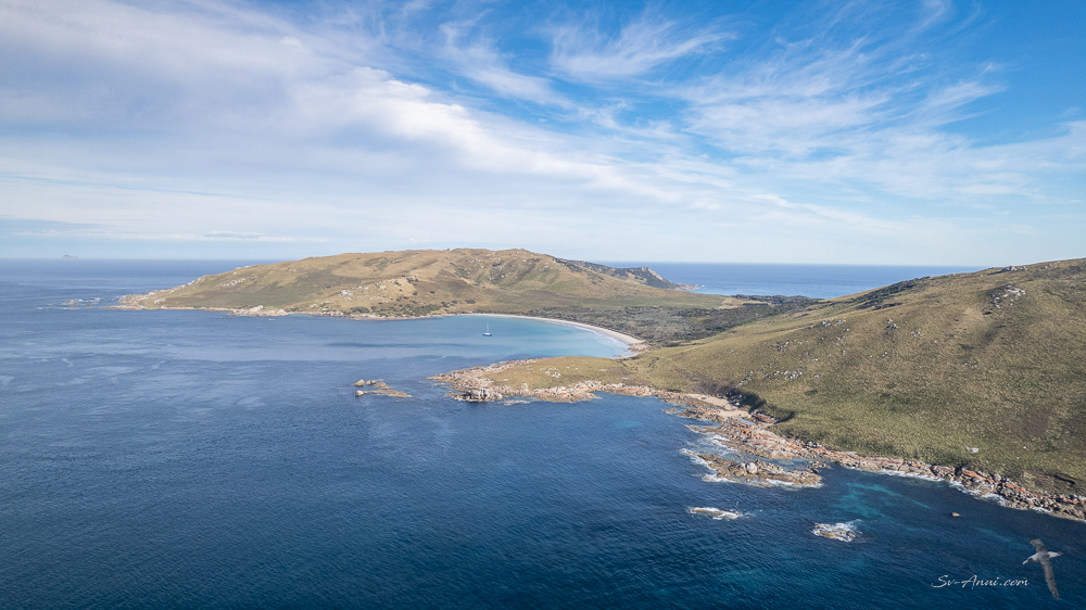 Aerial view of a scenic coastal landscape featuring a sandy beach, rocky shores, and green hills under a blue sky with scattered clouds.