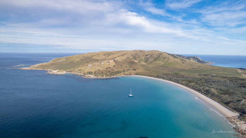 Aerial view of a coastal landscape featuring a sandy beach, clear blue waters, and a green hillside under a partly cloudy sky. A small sailboat is anchored in the bay.