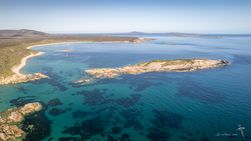 Aerial view of a tranquil coastal scene featuring clear turquoise waters, rocky formations, and a sandy beach bordered by lush greenery.