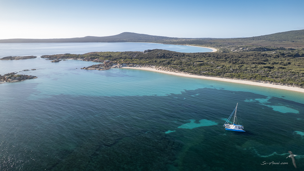 Aerial view of a serene coastal landscape featuring crystal-clear turquoise waters, sandy beaches, and a sailboat anchored near rocky shores.