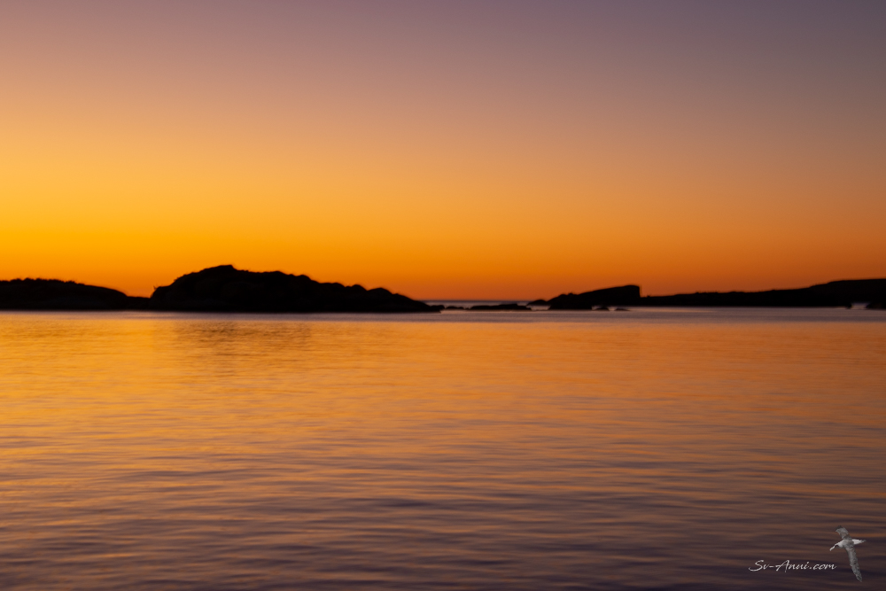 Sunset over a calm sea with silhouettes of rocks in the foreground and a gradient sky with orange and purple hues.