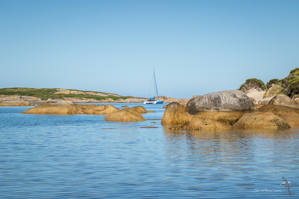 A serene coastal scene featuring smooth, large rocks in shallow water, with a sailboat anchored in the background and clear blue skies above.