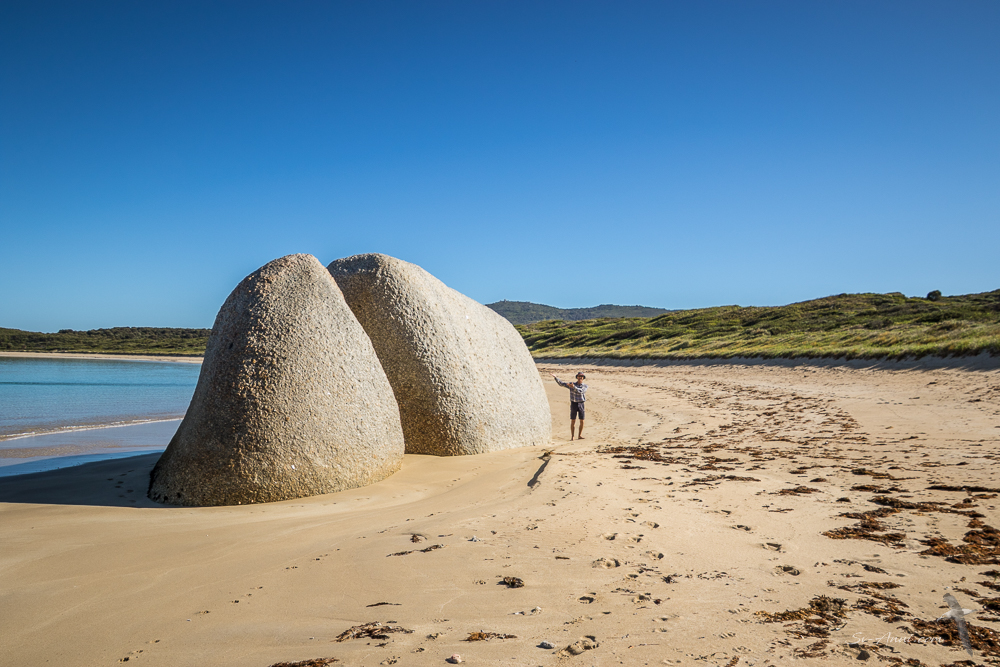 A person standing next to two large, smooth boulders on a sandy beach with a clear blue sky in the background.
