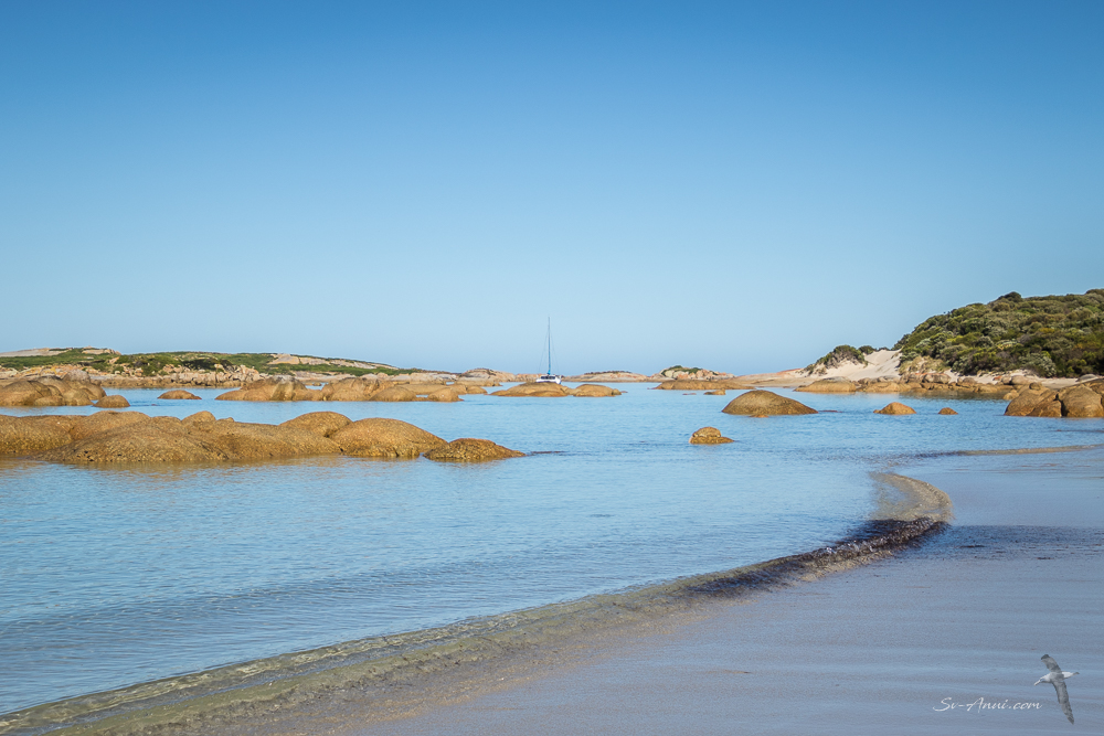 A serene coastal scene featuring smooth rocks scattered across shallow waters under a clear blue sky, with a sailboat visible in the distance.